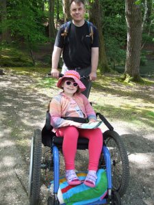 Annabelle in wheelchair in the forest. A man pushing the wheel chair. Both are smiling.